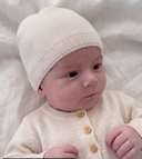 A close-up of a baby wearing a cream-colored knit beanie and matching cream cardigan with wooden buttons, lying on a soft white fabric background.