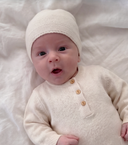 A baby lying on a white textured blanket, wearing a cream-colored knit beanie and a matching long-sleeve henley cardigan with three wooden buttons.