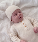A baby lying on a soft white blanket, wearing a cream-colored cashmere beanie and matching long-sleeve cashmere romper with wooden buttons.