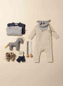 Flat lay of baby gift set including cream romper, knit booties, socks, toy horse, and matching checkered and solid pacifier straps