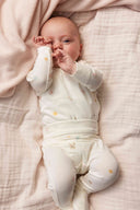 Baby lying down wearing cream kimono onesie with tiny toy print, rubbing eye with hand