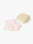 Pale pink kimono style wrap romper and hat folded next to an open beige gift box with white ribbon