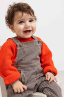 Baby sitting wearing brown and cream striped overalls over orange sweater, studio portrait