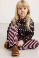 Girl sitting wearing dark brown knit sweater with pink heart stripe pattern and matching striped pants, brown boots