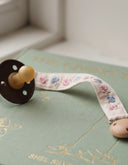 A dark brown pacifier with a yellow nipple rests on a sage green hardcover book with gold foil illustrations. A cream-colored fabric pacifier strap with a pink and blue floral print and a wooden bead end is draped across the book.