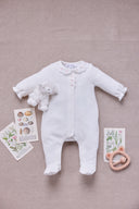 Flat lay of white sleepsuit with scalloped floral embroidered collar styled with plush lamb toy, wooden teether, and botanical postcards on linen background