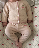 Baby wearing soft white pointelle knit cardigan and pants with strawberry embroidery and pink scalloped trim, lying on apple print blanket