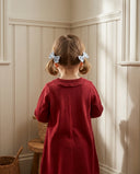 Girl wearing cherry red knit dress with ruffled collar, back view with floral hair bows, standing in paneled hallway