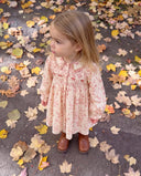A young girl with blonde hair stands outdoors on a ground covered in fallen autumn leaves, wearing a long-sleeve smocked dress in a light pink and white floral pattern with brown boots.