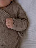 Close up of baby wearing taupe pointelle knit kimono jumpsuit with scalloped cuffs and tie fastening, hand grasping sleeve