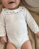 Close up of baby wearing ivory bodysuit with ruffled collar and blue floral smocking, on faux fur blanket
