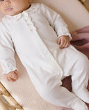 Baby lying in bassinet wearing white sleepsuit with scalloped floral embroidered collar, pink blanket in background