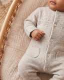 Baby wearing beige melange wool cashmere romper with button placket and patch pockets, lying in a woven bassinet