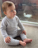 Baby sitting on the floor in light grey cashmere sweater and pants set, watching a duck through a rain streaked window