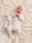 Baby wearing cream teddy bear print sleepsuit and knit bonnet, lying on cream blanket with white bunny lovey