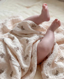 A close-up of a baby's bare legs wrapped in a soft cream muslin blanket with a brown teddy bear pattern, lying on a matching blanket.