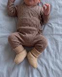 Baby wearing brown knit wrap romper and cream wool booties, lying on striped sheet