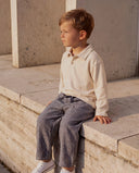 Boy sitting on a stone ledge wearing cream long sleeve polo with grey pinstripe trousers, white sneakers
