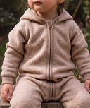 A close-up of a toddler wearing a cozy, light brown zip-up hooded sweatshirt and matching pants, sitting outdoors on a wooden surface with a blurred natural background.